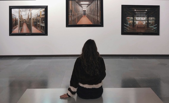 Eine Person sitzt vor einer Museumswand und betrachtet mehrere gerahmte Fotografien von Innenräumen.