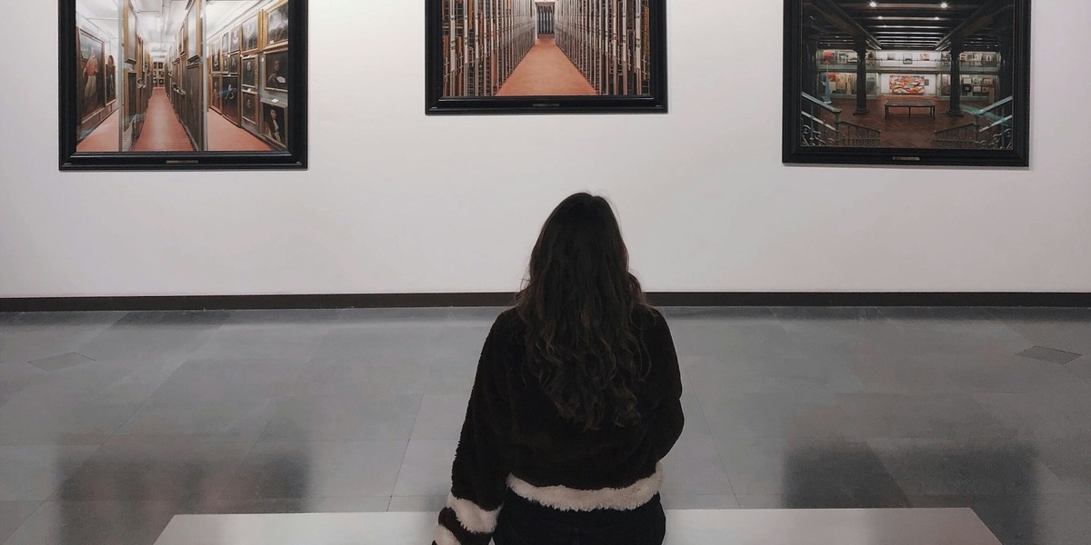 Eine Person sitzt vor einer Museumswand und betrachtet mehrere gerahmte Fotografien von Innenräumen.