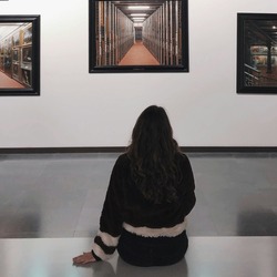 Eine Person sitzt vor einer Museumswand und betrachtet mehrere gerahmte Fotografien von Innenräumen.