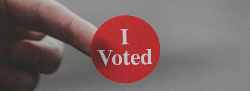 Eine Hand mit einem Aufkleber am Finger. Auf dem runden roten Sticker steht „I Voted“ (englisch für: „Ich habe gewählt“).