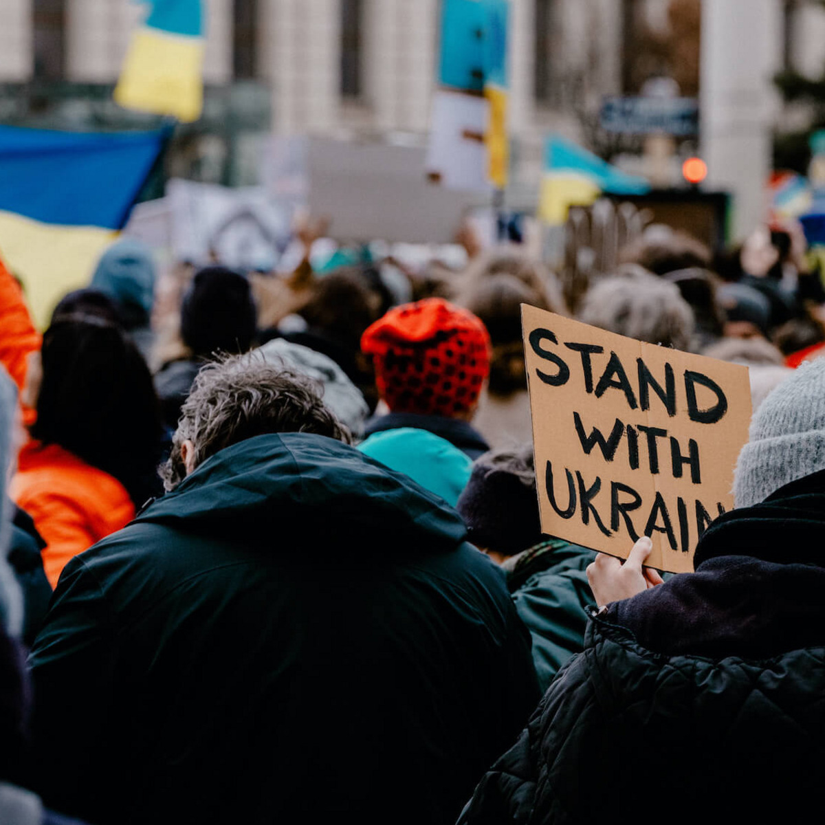 Viele Menschen stehen auf der Straße, einige von ihnen schwenken ukrainische Flaggen. Ein Mensch hält ein handgeschriebenes Schild mit der Aufschrift “Stand with Ukraine“ hoch.