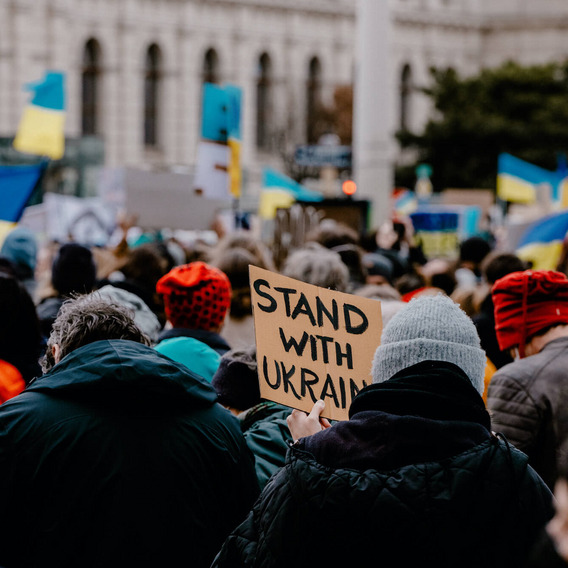 Viele Menschen stehen auf der Straße, einige von ihnen schwenken ukrainische Flaggen. Ein Mensch hält ein handgeschriebenes Schild mit der Aufschrift “Stand with Ukraine“ hoch.