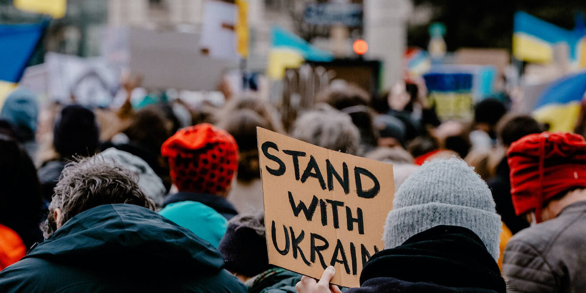 Viele Menschen stehen auf der Straße, einige von ihnen schwenken ukrainische Flaggen. Ein Mensch hält ein handgeschriebenes Schild mit der Aufschrift “Stand with Ukraine“ hoch.