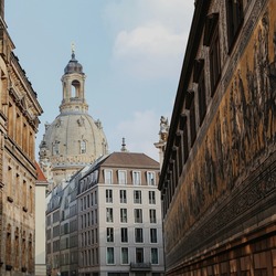Blick durch eine enge Altstadtgasse in Dresden auf die barocke Kuppel der Frauenkirche, flankiert von historischen Sandsteingebäuden. Rechts ist die Fürstenzugmauer mit ihrem berühmten Porzellanbild-Fries zu sehen.