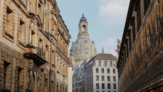Blick durch eine enge Altstadtgasse in Dresden auf die barocke Kuppel der Frauenkirche, flankiert von historischen Sandsteingebäuden. Rechts ist die Fürstenzugmauer mit ihrem berühmten Porzellanbild-Fries zu sehen.