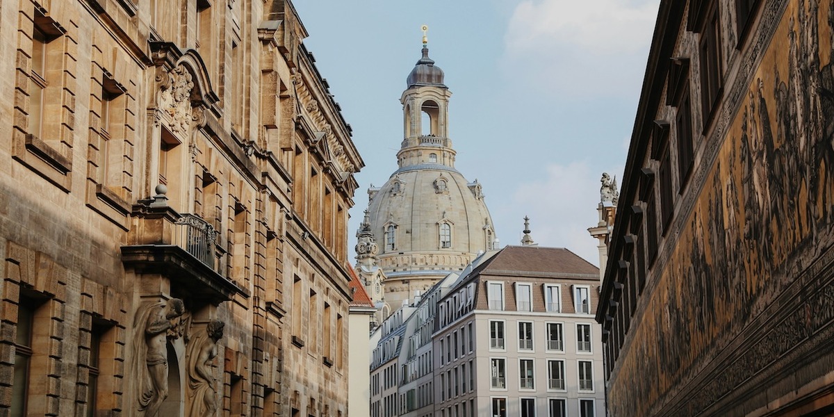 Blick durch eine enge Altstadtgasse in Dresden auf die barocke Kuppel der Frauenkirche, flankiert von historischen Sandsteingebäuden. Rechts ist die Fürstenzugmauer mit ihrem berühmten Porzellanbild-Fries zu sehen.