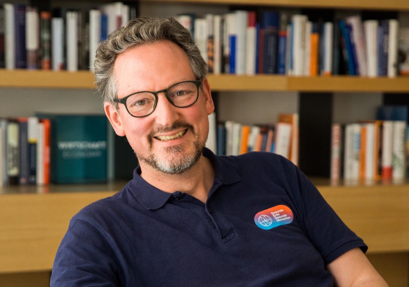 Eckart von Hirschhausen sitzt lächelnd vor einem Bücherregal. Er trägt eine Brille und ein dunkelblaues Polohemd mit einem kleinen Logo auf der linken Brust. Im Hintergrund sind zahlreiche Bücher zu sehen, die ordentlich in Regalen stehen. Von Hirschhausen wirkt freundlich und entspannt. Die Szene findet wahrscheinlich in einem Büro oder einer Bibliothek statt.