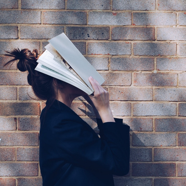 Ein Mädchen steht vor einer Backsteinmauer und hält ein Buch vor ihren Kopf.