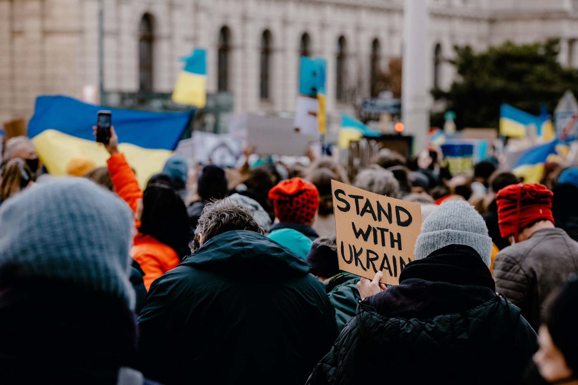 Viele Menschen stehen auf der Straße, einige von ihnen schwenken ukrainische Flaggen. Ein Mensch hält ein handgeschriebenes Schild mit der Aufschrift “Stand with Ukraine“ hoch.