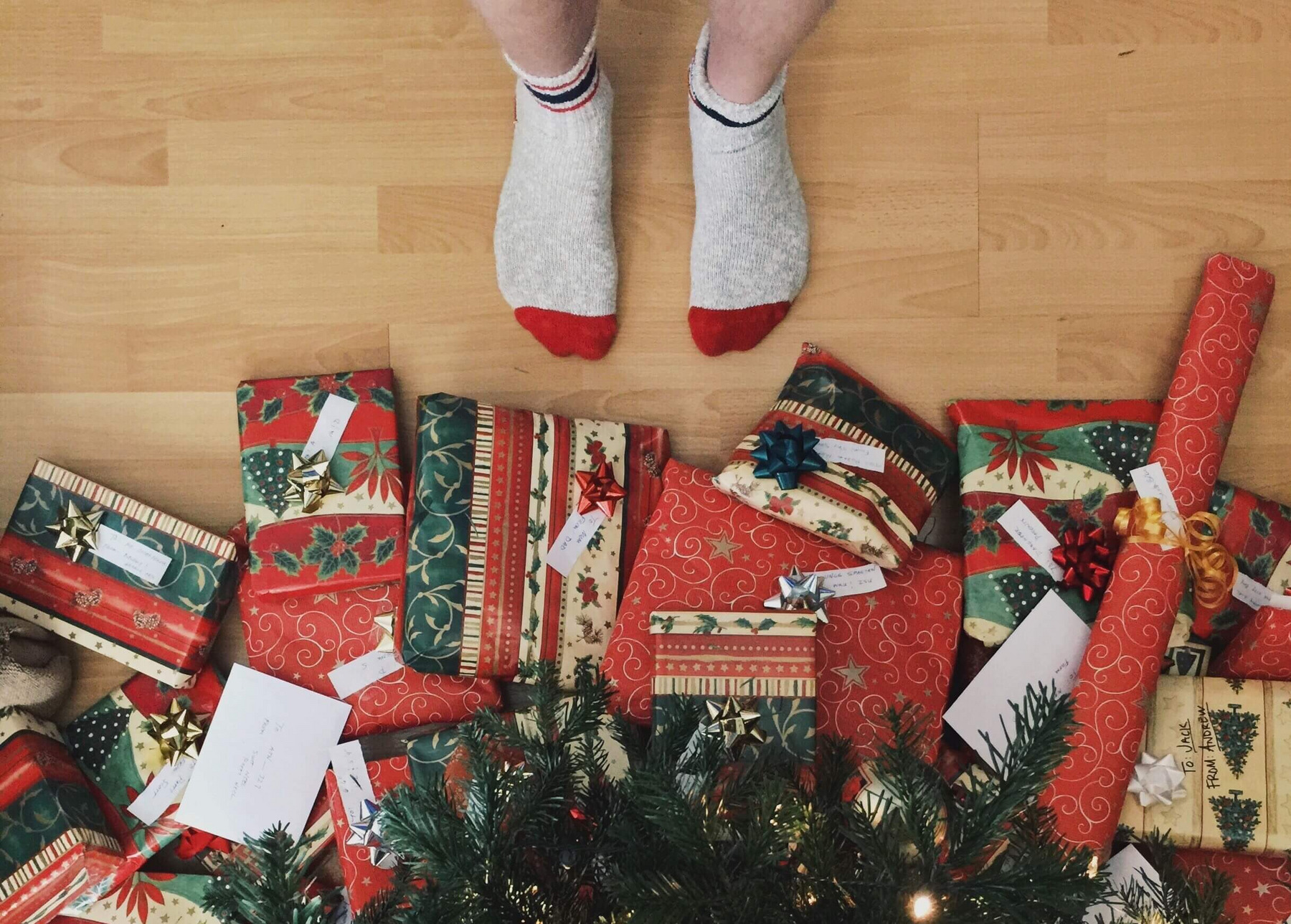 Eine Person in Socken steht vor einem Weihnachtsbaum, unter dem viele bunt eingepackte Geschenke liegen