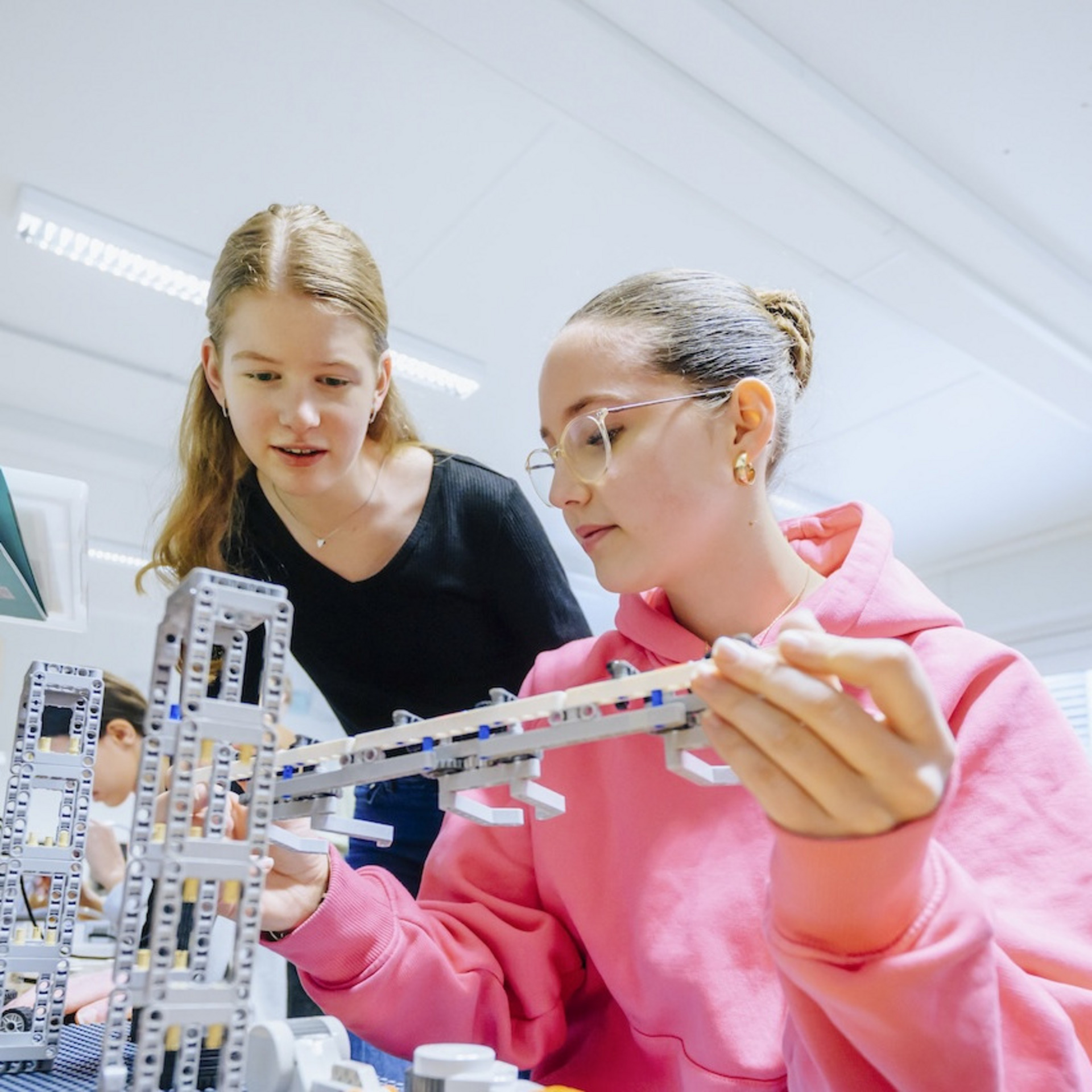 Zwei Schülerinnen arbeiten gemeinsam an einem Robotik-Projekt mit LEGO-Bausteinen in einem Klassenzimmer. Die Schülerin in der rosa Kapuzenjacke hält einen länglichen Bauteil in der Hand, während ihre Mitschülerin im schwarzen Shirt aufmerksam zuschaut.