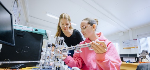 Zwei Schülerinnen arbeiten gemeinsam an einem Robotik-Projekt mit LEGO-Bausteinen in einem Klassenzimmer. Die Schülerin in der rosa Kapuzenjacke hält einen länglichen Bauteil in der Hand, während ihre Mitschülerin im schwarzen Shirt aufmerksam zuschaut.