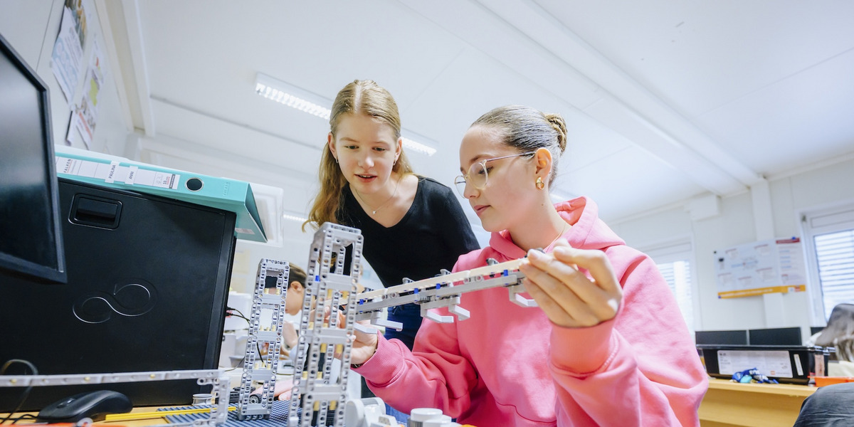 Zwei Schülerinnen arbeiten gemeinsam an einem Robotik-Projekt mit LEGO-Bausteinen in einem Klassenzimmer. Die Schülerin in der rosa Kapuzenjacke hält einen länglichen Bauteil in der Hand, während ihre Mitschülerin im schwarzen Shirt aufmerksam zuschaut.