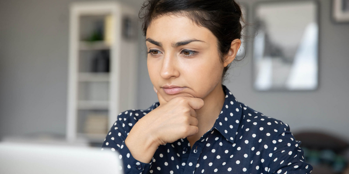 Eine Frau in gepunkteter Bluse sitzt vor einem Laptop und hat einen unsicheren, nachdenklichen Gesichtsausdruck.