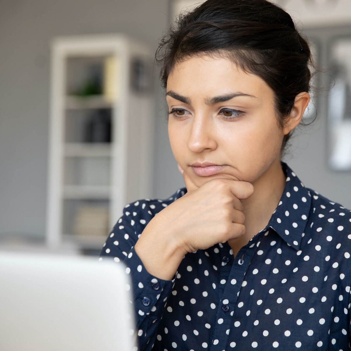 Eine Frau in gepunkteter Bluse sitzt vor einem Laptop und hat einen unsicheren, nachdenklichen Gesichtsausdruck.