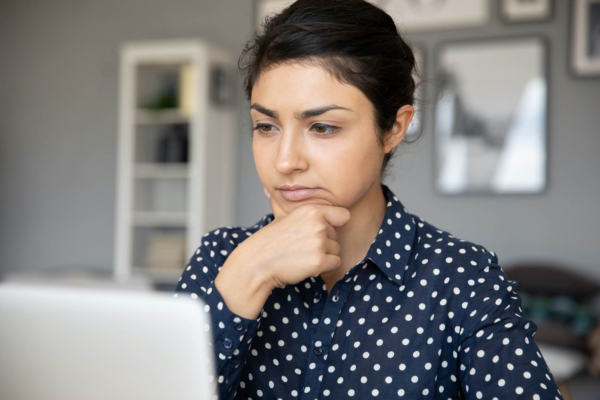 Eine Frau in gepunkteter Bluse sitzt vor einem Laptop und hat einen unsicheren, nachdenklichen Gesichtsausdruck.