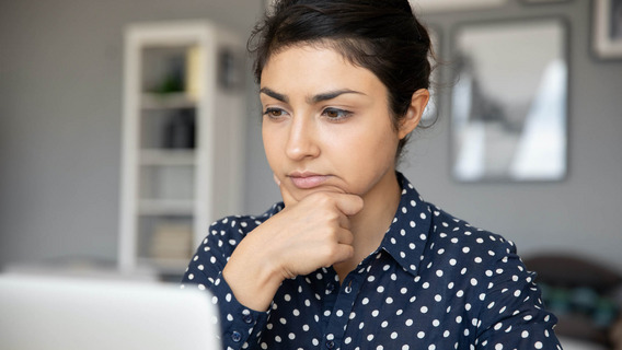 Eine Frau in gepunkteter Bluse sitzt vor einem Laptop und hat einen unsicheren, nachdenklichen Gesichtsausdruck.