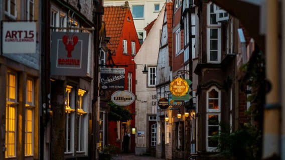 Eine schmale, kopfsteingepflasterte Gasse im Bremer Schnoor-Viertel, gesäumt von historischen Gebäuden mit leuchtenden Restaurant- und Geschäftsschildern bei Dämmerung.