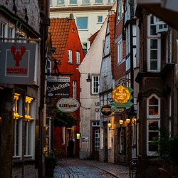 Eine schmale, kopfsteingepflasterte Gasse im Bremer Schnoor-Viertel, gesäumt von historischen Gebäuden mit leuchtenden Restaurant- und Geschäftsschildern bei Dämmerung.