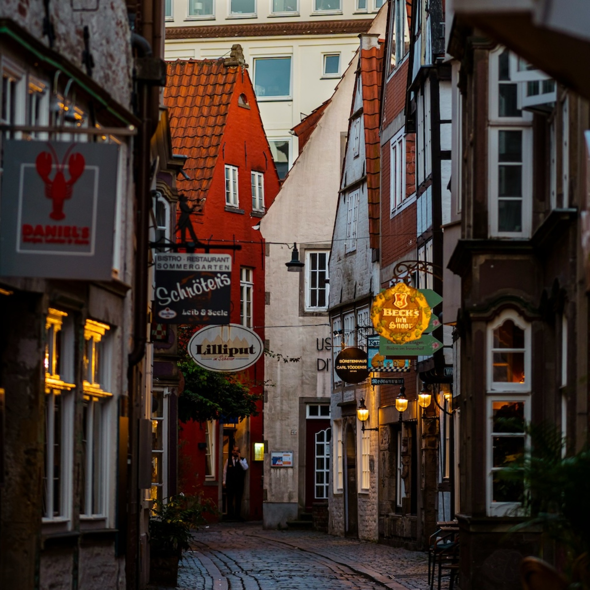 Eine schmale, kopfsteingepflasterte Gasse im Bremer Schnoor-Viertel, gesäumt von historischen Gebäuden mit leuchtenden Restaurant- und Geschäftsschildern bei Dämmerung.