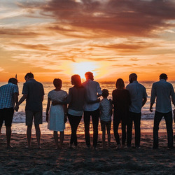 15 Menschen, aller Altersklassen und Geschlechter, stehen am Strand in einer Reihe und halten ihre Hände oder haben sich im Arm. Am Horizont geht die Sonne unter.