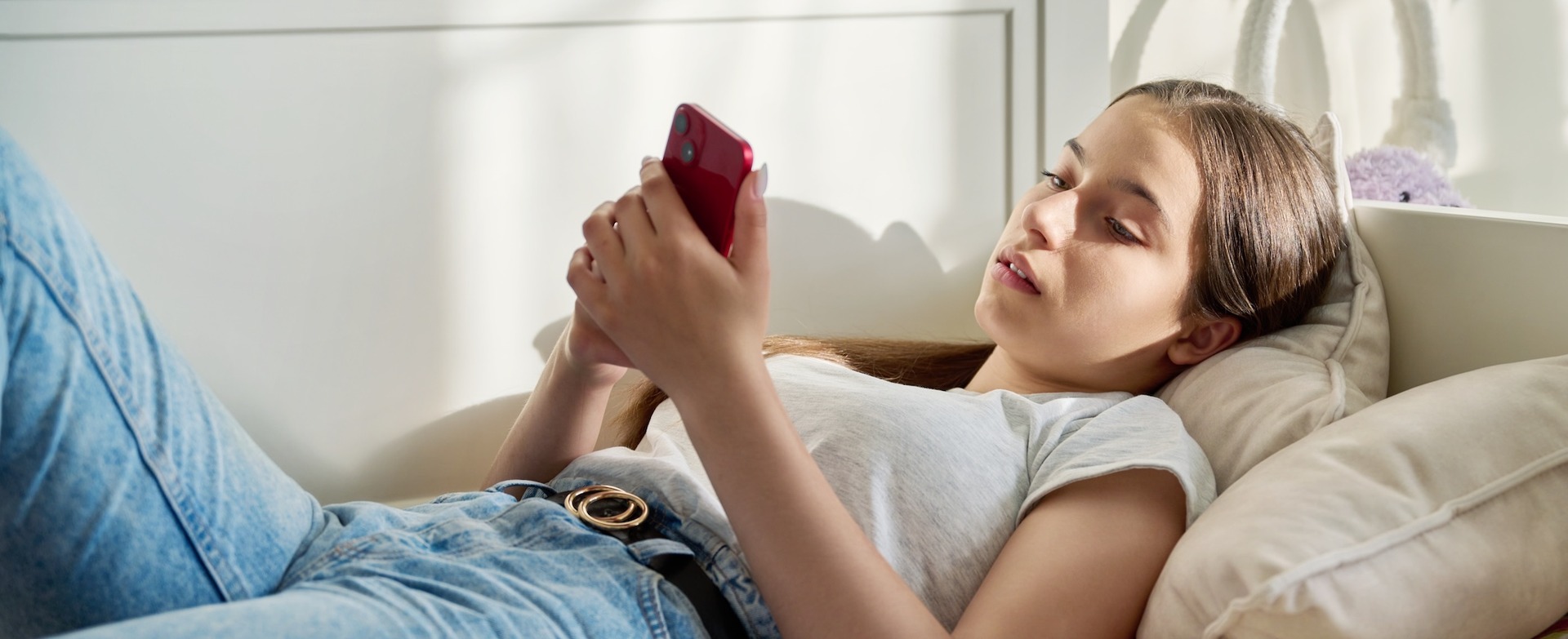 Jugendliche liegt entspannt auf einem Bett und schaut auf ihr rotes Smartphone. 