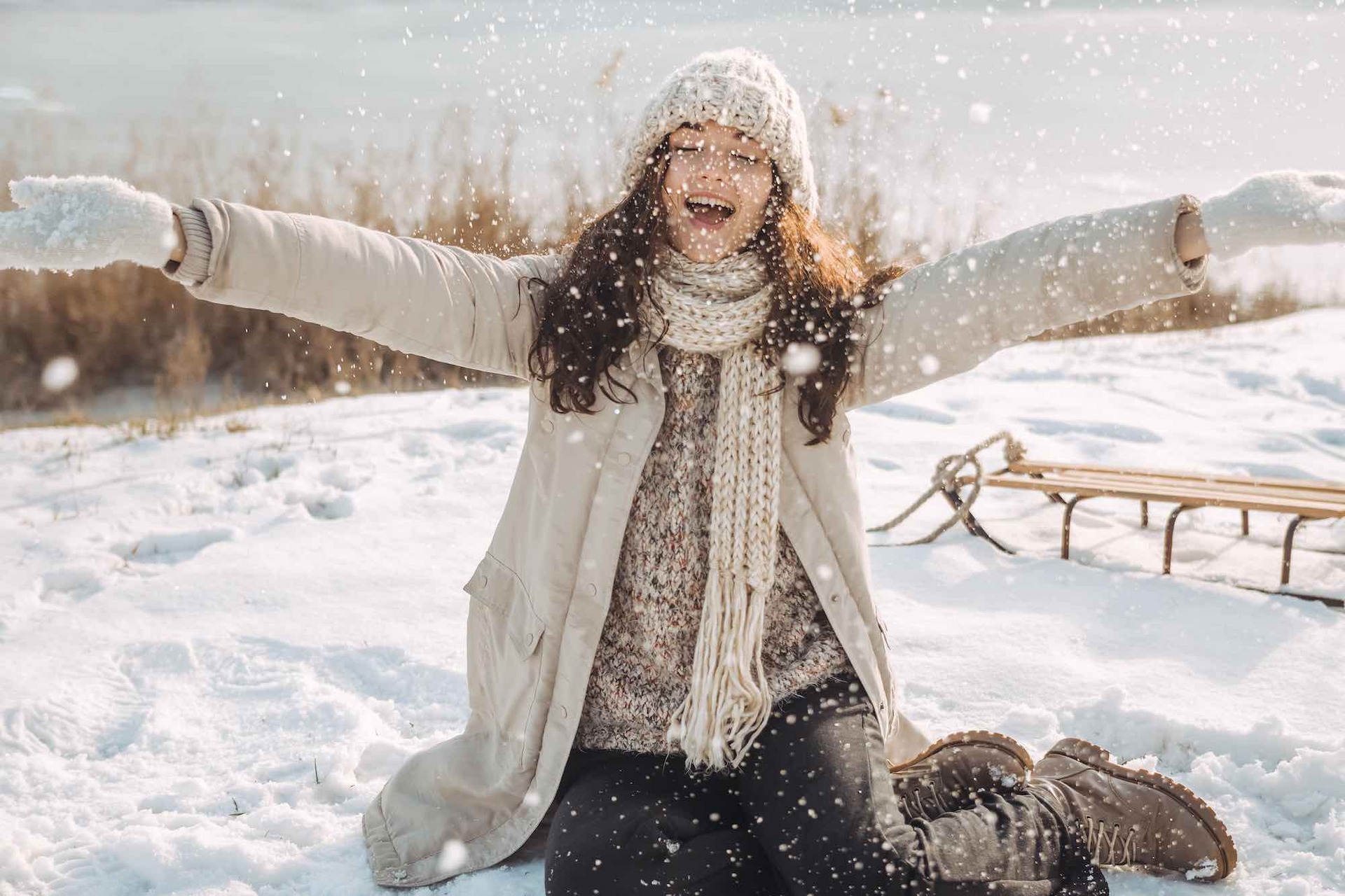 Eine junge, glückliche Frau sitzt in einer Schneelandschaft. Hinter ihr steht ein Schlitten auf dem Boden.