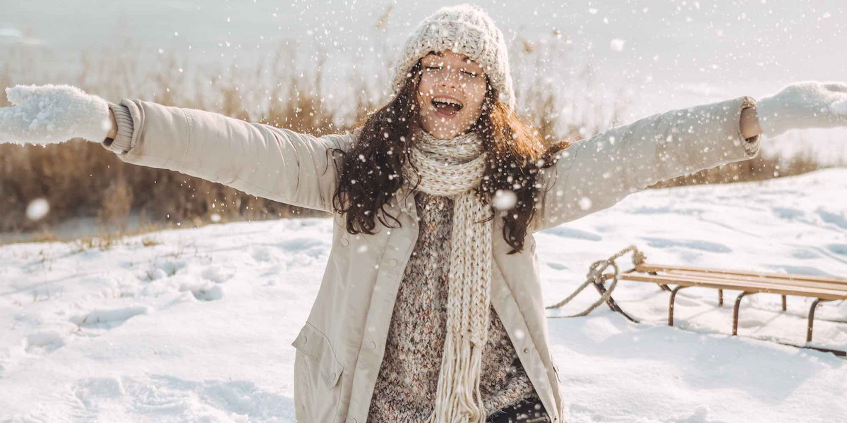 Eine junge, glückliche Frau sitzt in einer Schneelandschaft. Hinter ihr steht ein Schlitten auf dem Boden.