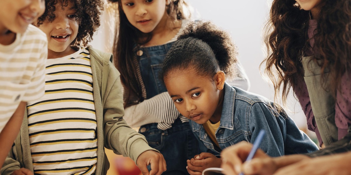 Eine Gruppe Kinder lehnt und steht an einem Tisch und beobachtet eine Person die etwas schreibt.