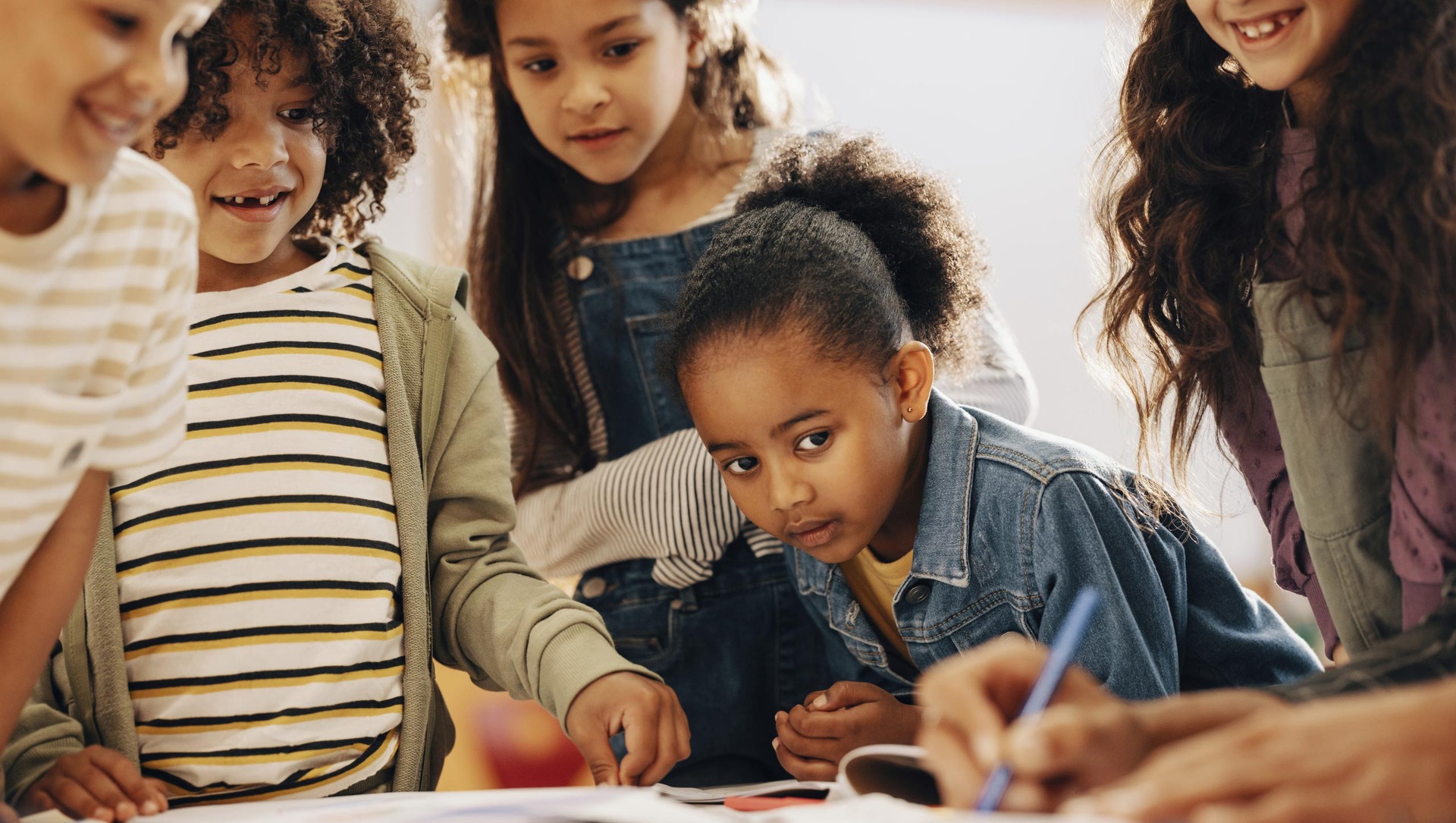 Eine Gruppe Kinder lehnt und steht an einem Tisch und beobachtet eine Person die etwas schreibt.