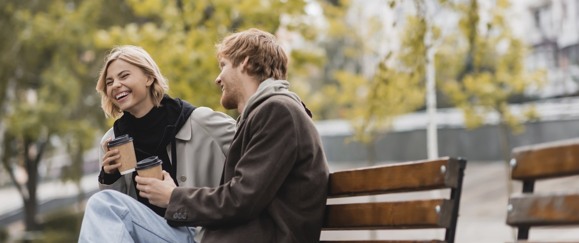 Zwei junge Menschen, eine Frau und ein Mann, sitzen entspannt auf einer Parkbank und unterhalten sich bei Kaffee. Im Hintergrund sind herbstliche Bäume und mehrstöckige Wohngebäude zu sehen.
