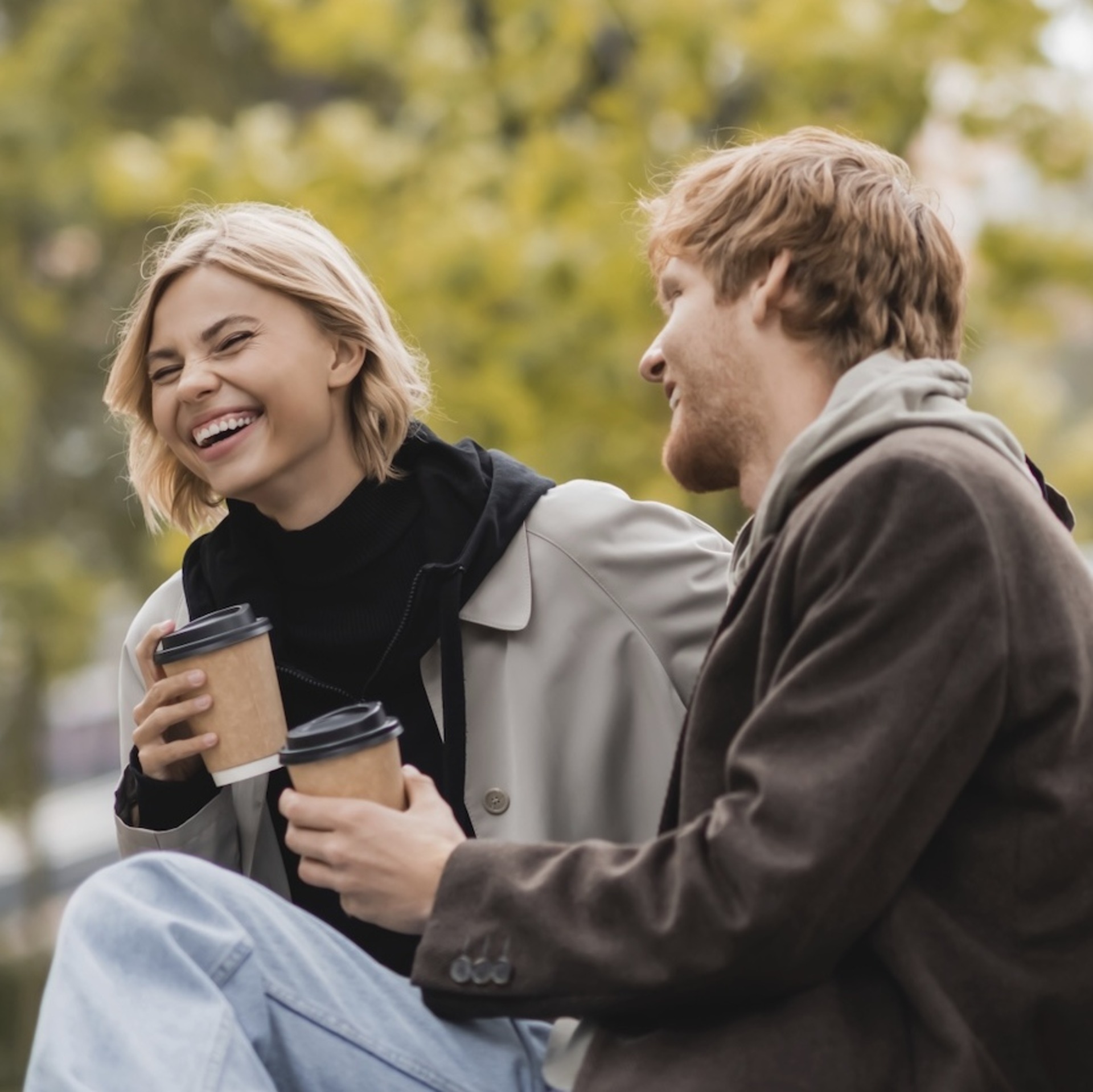 Zwei junge Menschen, eine Frau und ein Mann, sitzen entspannt auf einer Parkbank und unterhalten sich bei Kaffee. Im Hintergrund sind herbstliche Bäume und mehrstöckige Wohngebäude zu sehen.