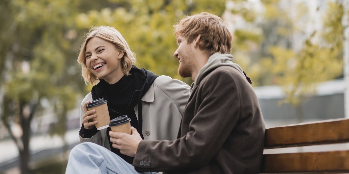 Zwei junge Menschen, eine Frau und ein Mann, sitzen entspannt auf einer Parkbank und unterhalten sich bei Kaffee. Im Hintergrund sind herbstliche Bäume und mehrstöckige Wohngebäude zu sehen.