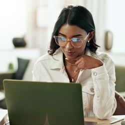 Eine Frau mit dunklen, schulterlangen Haaren und Brille sitzt an einem Tisch und arbeitet konzentriert an einem Laptop. Sie trägt eine helle Jacke und stützt nachdenklich ihre Hand ans Kinn. Auf dem Tisch liegen Notizen und ein Smartphone. Im Hintergrund ist ein heller, modern eingerichteter Raum mit Pflanzen und Sitzmöbeln zu sehen.