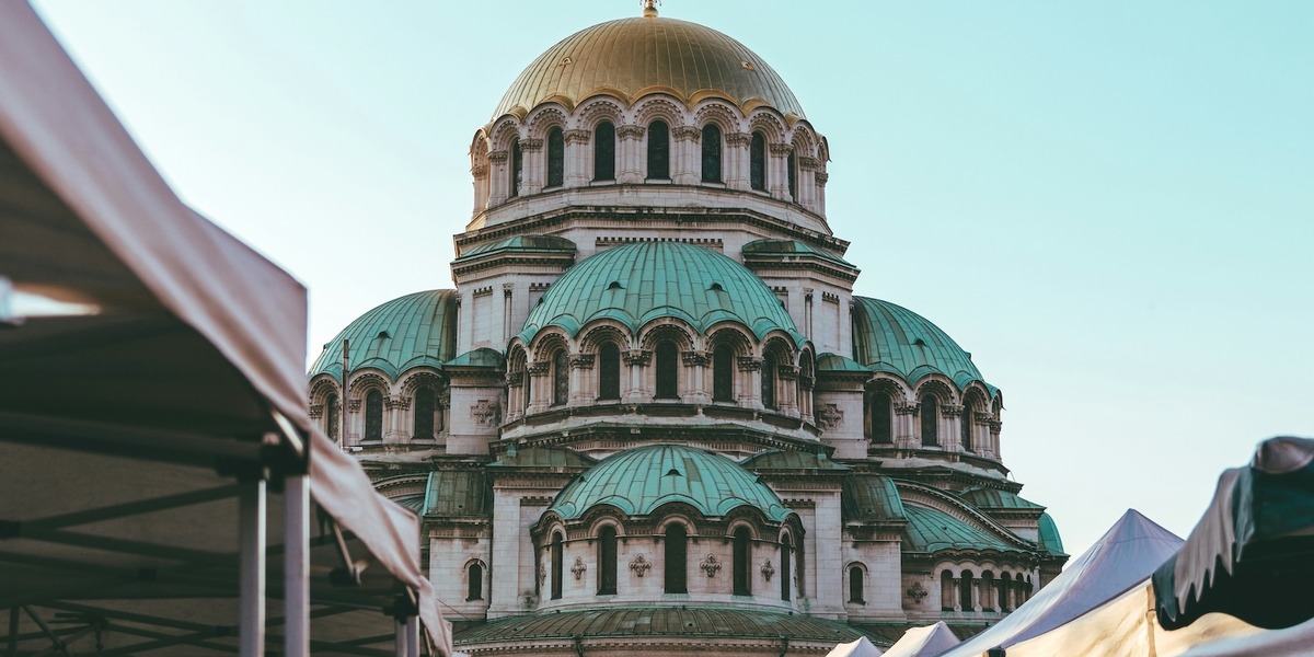 Eine Kuppelkirche mit grünen und goldenen Dächern. Im Vordergrund ragen die Dächer von Marktständen ins Bild.