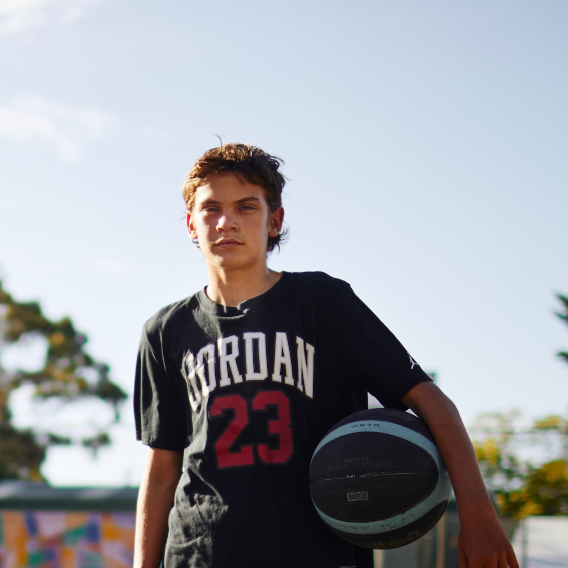 Ein Jugendlicher steht mit einem Basketball unter dem Arm auf einem Außenbasketballplatz. Im Hintergrund sind ein Basketballkorb, ein Zaun, Bäume und ein buntes Wandgemälde zu sehen.