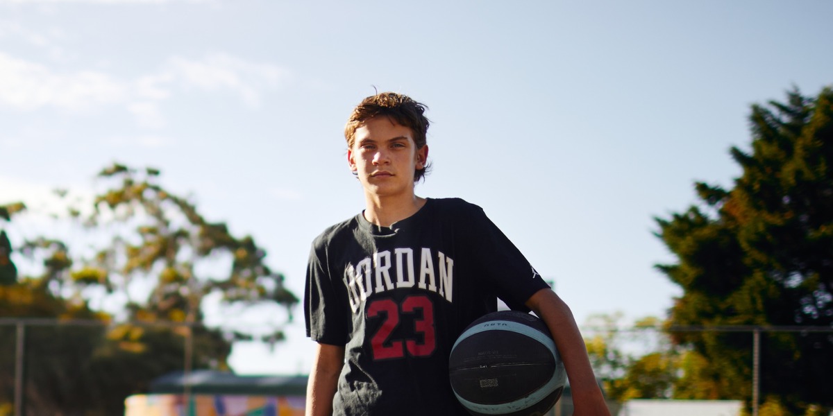 Ein Jugendlicher steht mit einem Basketball unter dem Arm auf einem Außenbasketballplatz. Im Hintergrund sind ein Basketballkorb, ein Zaun, Bäume und ein buntes Wandgemälde zu sehen.