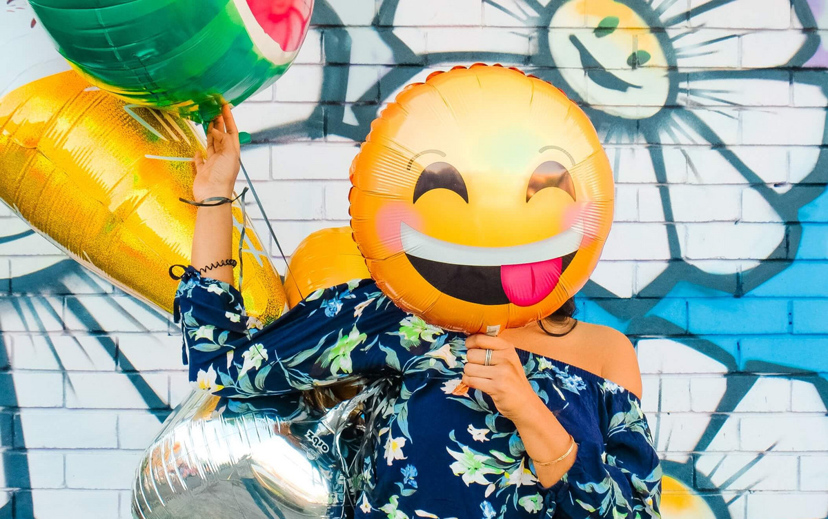 Ein großer Smiley-Luftballon verdeckt das Gesicht einer Person, die vor einer bunt bemalten Wand steht