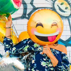 Ein großer Smiley-Luftballon verdeckt das Gesicht einer Person, die vor einer bunt bemalten Wand steht