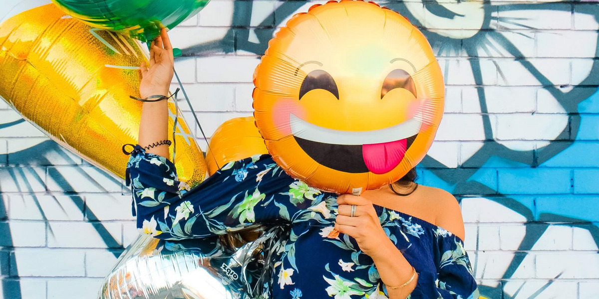 Ein großer Smiley-Luftballon verdeckt das Gesicht einer Person, die vor einer bunt bemalten Wand steht
