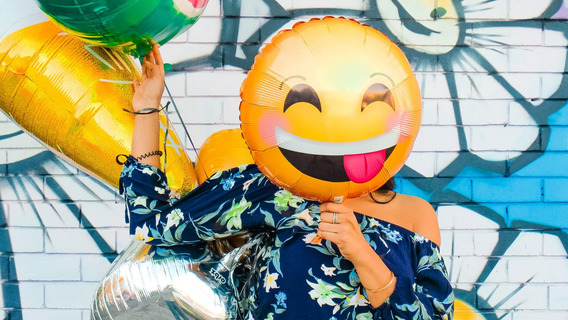 Ein großer Smiley-Luftballon verdeckt das Gesicht einer Person, die vor einer bunt bemalten Wand steht