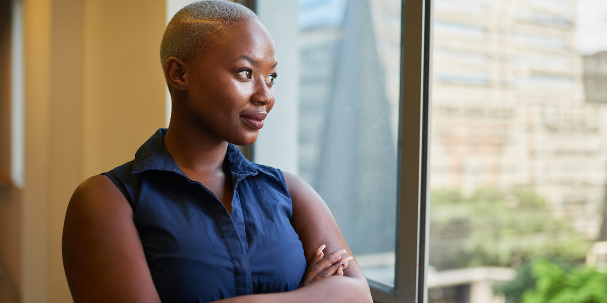 Eine Frau steht mit verschränkten Armen vor einem Fenster und schaut hinaus.