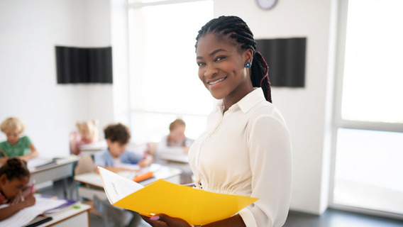 Eine lächelnde, Schwarze junge Frau hält eine gelbe, aufgeschlagene Mappe in der Hand. Im Hintergrund sind Kinder im Grundschulalter an Schultischen zu erkennen.