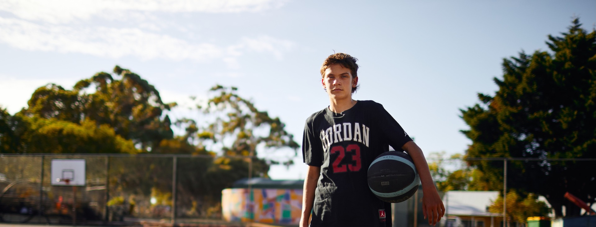 Ein Jugendlicher steht mit einem Basketball unter dem Arm auf einem Außenbasketballplatz. Im Hintergrund sind ein Basketballkorb, ein Zaun, Bäume und ein buntes Wandgemälde zu sehen.