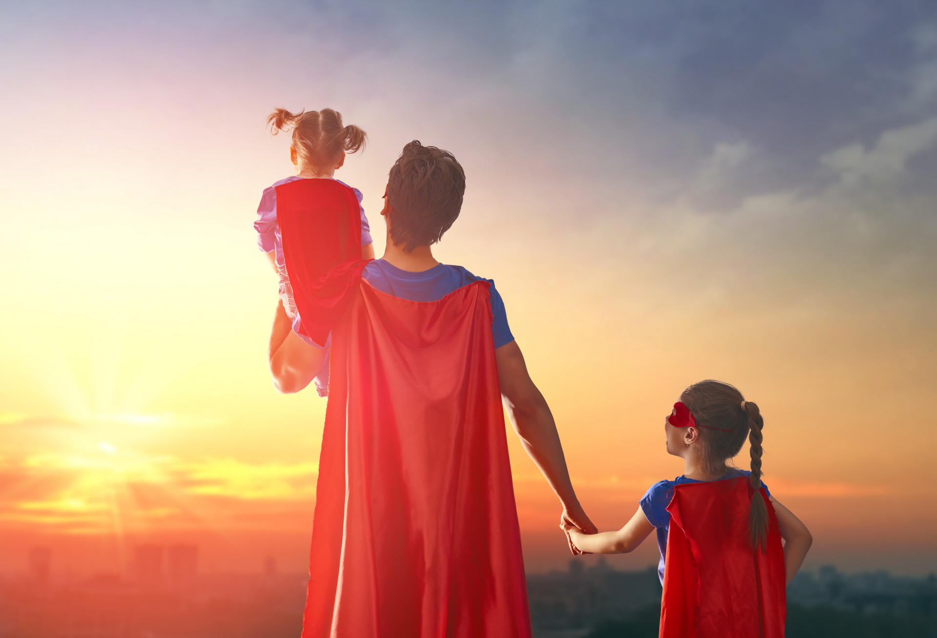 Ein als Superheld verkleideter Mann blickt mit Kindern in einen Sonnenuntergang