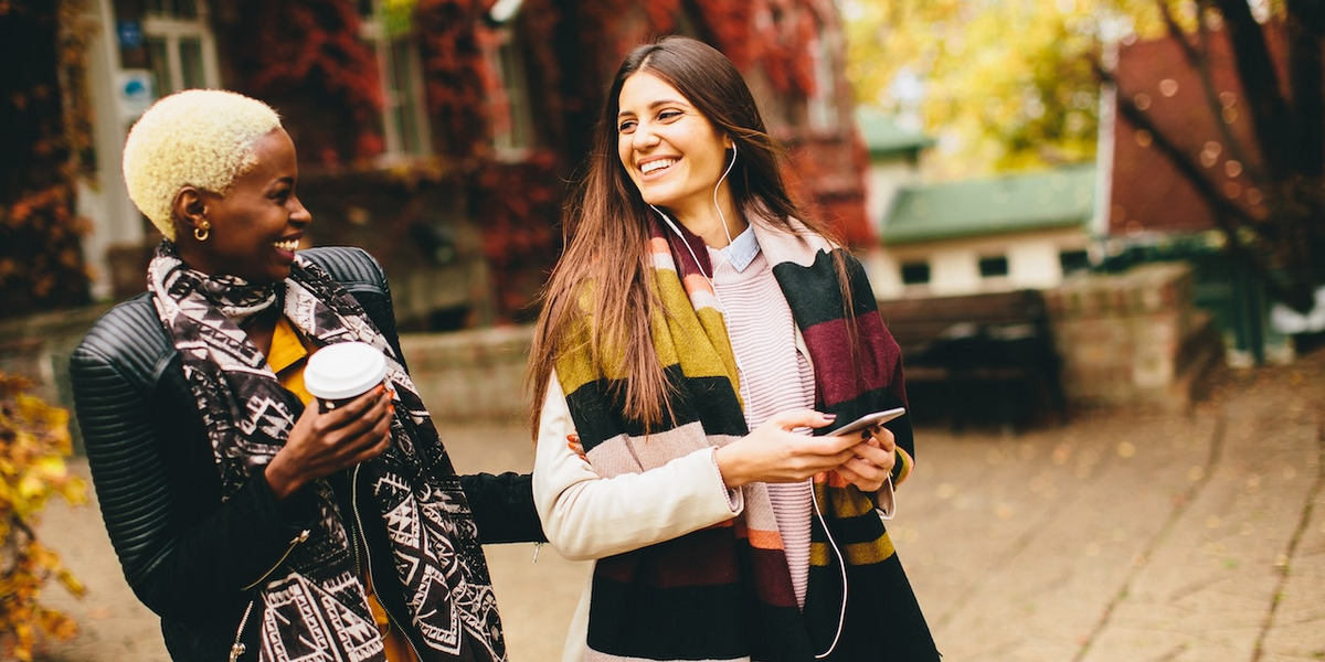 Zwei junge Frauen schlendern durch eine herbstliche Stadt und lachen miteinander.