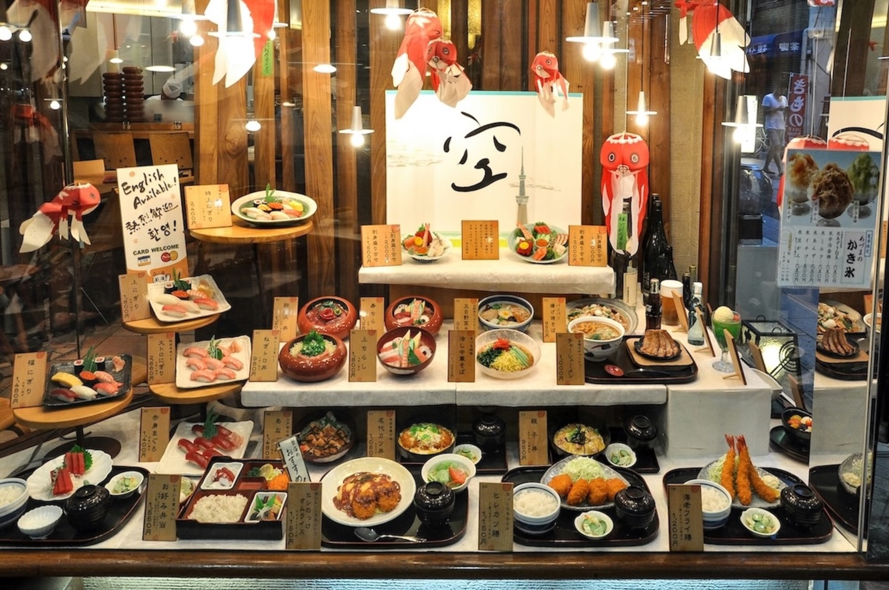 Restaurant-Schaufenster in Japan mit zahlreichen realistisch nachgebildeten Gerichten und japanischer Beschriftung, im Hintergrund eine Zeichnung.