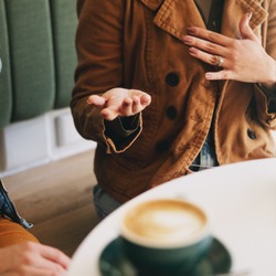 Zwei Personen sitzen an einem Tisch in einem Café. Vor ihnen stehen zwei Tassen mit Kaffee. Eine Person gestikuliert, während sie spricht.