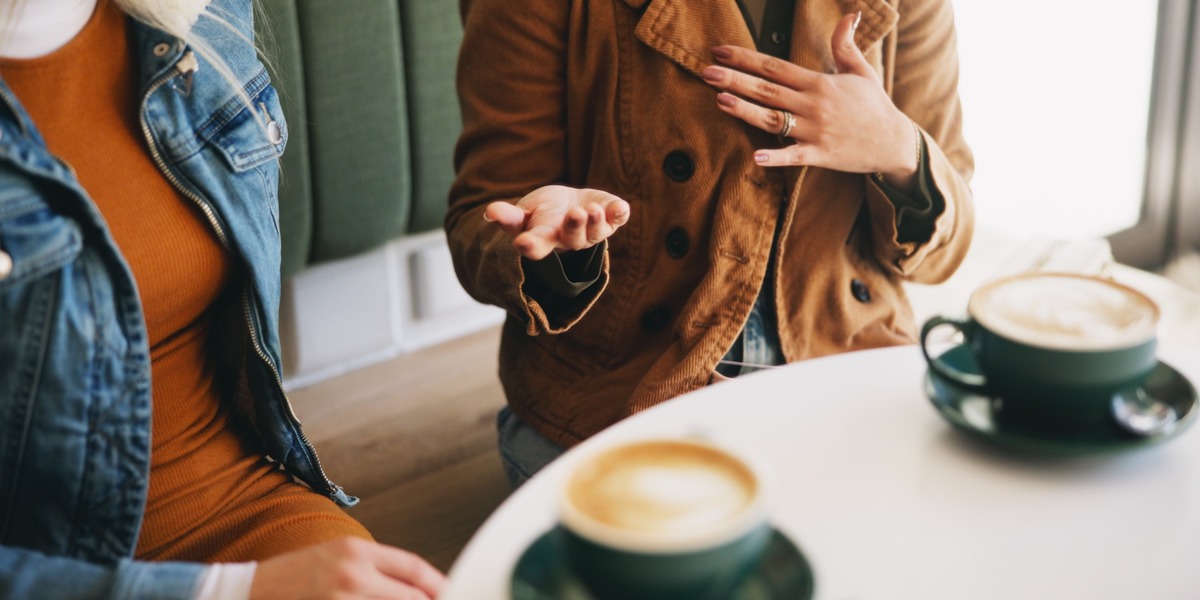 Zwei Personen sitzen an einem Tisch in einem Café. Vor ihnen stehen zwei Tassen mit Kaffee. Eine Person gestikuliert, während sie spricht.