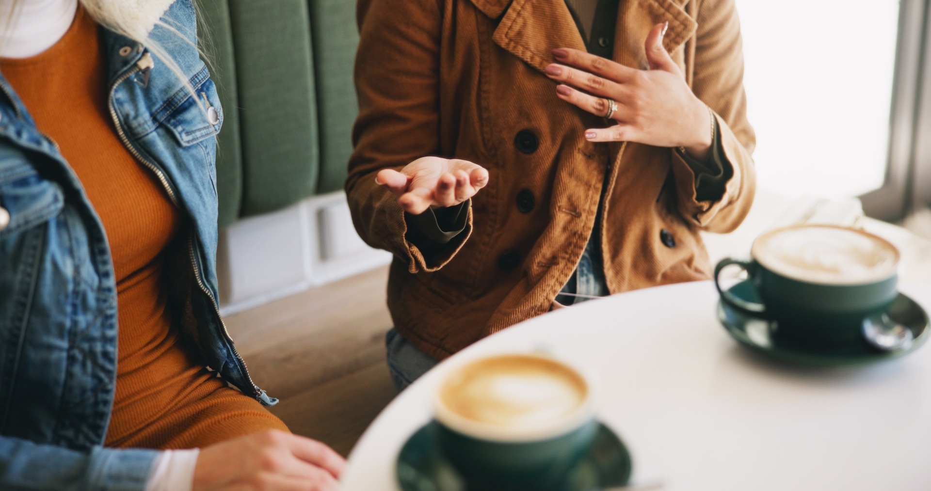 Zwei Personen sitzen an einem Tisch in einem Café. Vor ihnen stehen zwei Tassen mit Kaffee. Eine Person gestikuliert, während sie spricht.